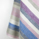 Yunga baby alpaca wool blanket – a soft, woolen textile displays diagonal pastel stripes in pink, blue, green, and purple hues. It hangs gently, offering a subtle texture in a neutral, indoor setting