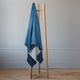 A wooden clothes rack supports a blue and gray fringed blanket draped over it. The setting is a minimalist room with light gray walls and herringbone wood flooring.