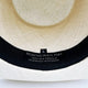 A cream-colored woven hat with a black inner band, labeled MONTECRISTI HAT, 100% PAJA TOQUILLA, HANDWOVEN IN ECUADOR, L. The hat's intricate texture is prominent.