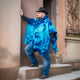 A person wearing a blue tie-dye poncho and black hat leans against a pillar on stone steps, with an ornate wall in the background.