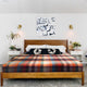 Anzu baby alpaca wool blanket – a wooden bed with a colorful plaid blanket and decorative pillows sits between two nightstands, featuring lamps and plants. Abstract artwork hangs above on the white wall