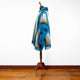 A mannequin displays a fringed, multi-shaded blue and grey poncho. The poncho drapes elegantly, set against a plain white wall and polished wooden floor.