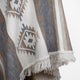 A fringed woven textile displays geometric patterns in beige, gray, and white, hanging against a plain background.