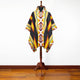 A colorful poncho with geometric patterns in red, yellow, and black hangs on a wooden mannequin. It features a hood and a buttoned neckline, set against a white background.