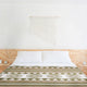 A neatly made bed with white pillows and a patterned green blanket sits in a minimalist room. A macramé wall hanging decorates the white wall above the wooden headboard.