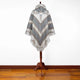 A poncho with geometric patterns hangs on a wooden stand. It features a hood and fringe trim, displayed against a plain white wall on a wooden floor.