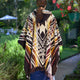 A person stands outdoors wearing a colorful poncho with geometric patterns. They face a path surrounded by greenery and flowers, with a table and chairs visible in the distance.