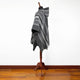 Mannequin displays a gray and white striped poncho with a hood, positioned on a wooden stand, against a plain white wall and hardwood floor.