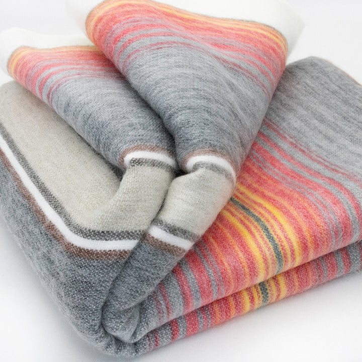 Ajilanga baby alpaca wool blanket – a folded, multicolored wool blanket with stripes in gray, red, orange, and white; resting on a white surface