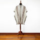 A cream-colored poncho with intricate brown geometric patterns is displayed on a wooden stand, set on a polished wooden floor against a plain white wall.