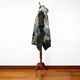 A hooded, tie-dye poncho is draped on a wooden mannequin stand, displaying shades of blue and brown. It is set against a white wall and wooden floor background.