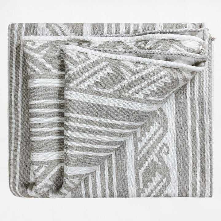 A folded blanket with intricate geometric patterns in grey and white, displaying intersecting lines and shapes, is laid flat against a plain background, highlighting its texture and design.