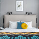 A bed is neatly made with a beige headboard, adorned with white, blue, and yellow pillows, under a framed heron picture. Patterned blanket covers part of the bed.