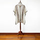 A poncho with intricate geometric patterns hangs on a wooden stand. The poncho features earthy tones and fringe detailing, set against a white wall and wooden floor.
