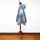 A light blue poncho with fringe detailing is draped on a wooden mannequin stand against a plain white background, set on a polished wooden floor.