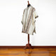 A poncho with intricate brown and black geometric patterns is displayed on a wooden mannequin stand, set against a plain white wall and wooden floor.