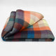 Anzu baby alpaca wool blanket – a folded, multi-colored plaid blanket featuring a pattern of squares in shades of orange, blue, green, and gray lies on a plain, neutral surface