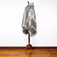 A patterned poncho draped on a wooden mannequin stand, featuring geometric designs with fringe edges, set against a plain white background and wooden floor.