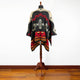 A poncho with geometric patterns in black, red, and beige is draped on a wooden mannequin stand. It stands in a room with a wooden floor and white wall.
