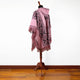 A pink, fringed poncho with black geometric patterns is draped on a wooden mannequin stand on a wooden floor, against a plain white background.