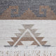A woven rug displays geometric patterns with brown and gray shapes. The design features stepped motifs against a textured backdrop, and white fringe hangs at the bottom edge.