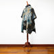 A colorful, abstract-patterned poncho with a hood is displayed on a wooden mannequin stand. It is in a room with polished hardwood flooring and a plain white wall.