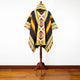 A vibrant, geometric-patterned poncho displays on a wooden stand. It has bold red, yellow, and black stripes with intricate designs, set against a plain white background and hardwood floor.