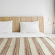 Quijos alpaca wool blanket – a neatly made bed with white pillows and a striped blanket sits against a wooden headboard, surrounded by a minimalist white room with a hanging light bulb