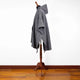 A gray, textured poncho drapes over a wooden mannequin stand, set against a white background and wooden floor, showcasing its hood and wide sleeves.