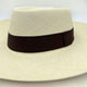A beige woven hat with a wide brim and a dark brown band sits on a neutral background, highlighting its elegant, classic design.