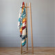 A wooden coat stand holds a colorful, geometric-patterned blanket, situated in a minimalist room with light gray walls and a parquet wood floor.