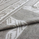 A folded, patterned fabric with tribal motifs in gray and white lies on a flat surface, displaying angular geometric designs and subtle texture in a close-up view.