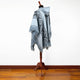 A light blue and gray patterned poncho with fringed edges is draped over a wooden stand in a room with hardwood flooring and plain white walls.