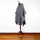 A gray, textured poncho drapes over a dark mannequin in a minimalist room with wooden flooring. The poncho has a loose, casual fit with pointed edges.