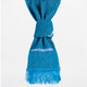 A blue woven scarf hangs downward, featuring a decorative band with geometric patterns near a section tied in a knot. It has fringed ends, against a plain background.