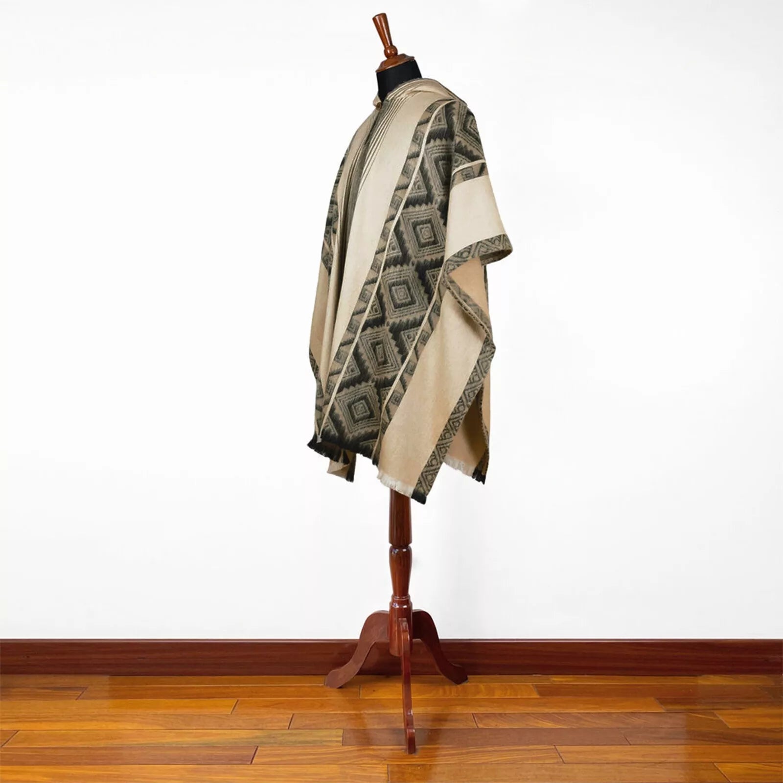 A beige poncho with geometric patterns drapes over a wooden mannequin stand. It's displayed on a polished wooden floor against a plain white wall.