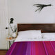 Chone baby alpaca wool blanket – bed with a wooden headboard displays white pillows and colorful blanket. A small table holds a cup, candle, and vase with greenery. Wall features black lamp and hanging foliage
