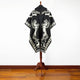 A black cloak with white horse designs hangs on a wooden stand against a white wall and wooden floor, showcasing intricate patterns and a fringe hem.