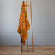 Putumayo baby alpaca wool blanket – a striped, orange and green blanket hangs on a simple wooden stand with hangers, positioned in a room with light-colored walls and parquet flooring, creating a minimalist aesthetic