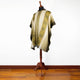 A poncho with geometric patterns drapes on a wooden mannequin stand, set against a white wall in a room with a wooden floor.