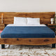 A wooden bed frame supports the neatly made bed with a blue blanket and white pillows. Positioned in a minimal bedroom, it features candles, books, and a plant on bedside tables.