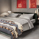 Bed adorned with gray linens and a patterned blanket, situated in a modern bedroom. Two red abstract paintings hang above, while a lamp and vases decorate the bedside tables.