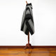 A black and white patterned poncho is displayed on a wooden stand. The poncho features geometric designs and fringe edges, situated in a room with a wooden floor and white wall.