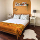Putumayo baby alpaca wool blanket – a neatly made bed with patterned pillows and a striped blanket sits against a mustard accent wall with good night text. It is flanked by white bedside tables and decor