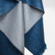 A thick, soft blue and gray fabric with fringed edges hangs against a plain background, showing its texture and layered folds.