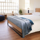 A neatly made bed with a light gray blanket is positioned in a modern bedroom. It features wooden furniture and a large window with a grid design, allowing natural light in.