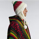 Person wears a red, green, and cream knitted winter hat with ear flaps and pom-poms, paired with a colorful patterned poncho against a plain gray background.