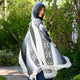 Palora lightweight alpaca wool hooded poncho – a person wrapped in a fringed, patterned poncho stands outdoors, turning slightly with a smile. They are surrounded by green foliage and a garden path under sunlight