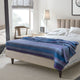 Morona baby alpaca wool blanket – a neatly made bed with a blue-striped blanket sits in a bright room. Nearby, a lamp and framed photo rest on a nightstand beside a wooden chair