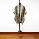 A geometric-patterned poncho drapes over a mannequin stand, set against a plain white wall and wooden floor.
